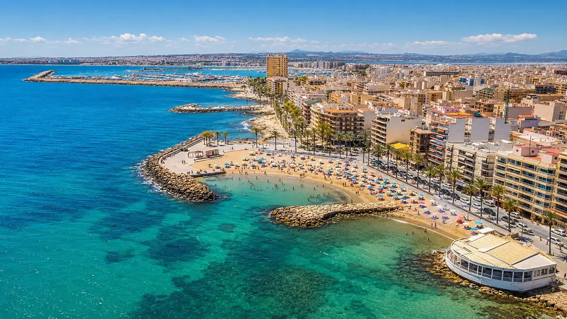 Aerial view of the Torrevieja seafront — a sandy town beach between rock breakwaters, the palm-lined promenade and the apartment blocks of the centre, with the marina beyond